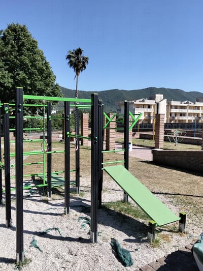 The image depicts an outdoor exercise area in Sessa Aurunca, Italy. The scene includes a jungle gym-like structure with black and green bars, gymnastics rings hanging from chains, and a green inclined sit-up board. The ground is covered with light-colored gravel interspersed with patches of green netting.

In the background, there are brick pillars, low brick walls, and additional exercise equipment. A building with beige walls and multiple windows is visible behind the exercise area, and a palm tree stands in the distance. The backdrop also includes a clear blue sky and low-lying mountains. The lighting indicates it is daytime. There are no people present in the image.