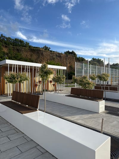 The image captures a modern outdoor public space or promenade on a bright, sunny day in Taman, Russia. In the foreground, clean, white rectangular planter boxes or low walls define the area, contrasting with the grey paved ground, which includes sections of intricate circular grates.

Integrated behind these white structures are several seating areas featuring slatted brown wooden benches. Adjacent to these benches, small, ornamental trees or shrubs with variegated green and light-colored foliage are planted in white rectangular planters. Further in the background, a tall, white metal fence extends horizontally, separating the manicured area from a natural, steep hillside. This hillside is characterized by dry, light brown earth interspersed with patches of green trees and shrubs.

The upper portion of the scene is dominated by a clear, vibrant blue sky dotted with scattered wispy white clouds, indicative of fine weather during daytime, likely morning or early afternoon. The scene is devoid of people, presenting a tranquil and well-maintained environment. No discernible text is visible within the frame.