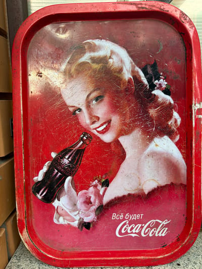 A vintage Coca-Cola tin tray is displayed horizontally. The tray features a distressed red background with the image of a blonde woman holding a bottle of Coca-Cola. She is smiling and looking at the viewer. There are pink flowers near her hand. In the lower right corner of the tray, Cyrillic text reads "Всё будет" (Everything will be) above the Coca-Cola logo. The tray exhibits significant wear, including scratches, scuffs, and rust, particularly along the edges and in the upper portion of the image. The tray is placed on a grey stone surface with a blurred brick wall in the background, suggesting it is outdoors or in a rustic setting. It is not possible to determine the specific location within the city of Каменка, Russia, from the visual information provided.