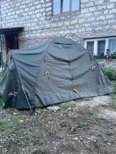 In the city of Teresva, Ukraine, a dark green tent is set up on a patch of ground with scattered rocks and sparse vegetation in front of a weathered brick building. The tent is a dome or tunnel style, with yellow guy ropes attached to the sides and a visible brand logo that appears to read "TERR INC." with "ZETA" underneath. The building features a dark-colored door and frame on the left, and a window with multiple panes and flower pots on the right. A yellow pipe runs horizontally across the building's facade, and there are also exposed bricks near the bottom of the wall. The ground is uneven, a mix of dirt, pebbles, and some patches of mud, with various weeds and grasses growing throughout.