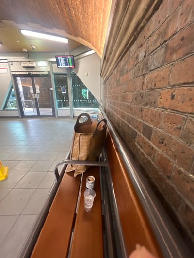 The image shows a view of a waiting area at Chester railway station, United Kingdom. The scene is captured from a seated perspective, looking towards a brick wall and a seating bench.
The main subjects in the foreground are a brown woven tote bag placed on the bench, a partially consumed can of a beverage, and a clear plastic cup with some liquid inside. The bench itself is made of polished wooden planks with metal supports. A portion of a yellow hazard cone is visible on the tiled floor to the left of the bench.
In the background, there is a departure board displaying train information, indicating the location as Chester. To the left, glass doors lead to an exterior area. The architecture includes a curved ceiling, likely brickwork, and fluorescent lighting. The overall environment appears to be an indoor waiting area, possibly a station concourse or platform area. The time of day seems to be daytime, given the lighting conditions. There are no other people clearly visible in the frame, although there might be a hint of a hand in the bottom right corner.