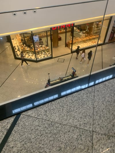 This image captures an overhead view of the ground floor within a multi-level shopping mall, specifically identified as **Parque Shopping Bahia in Lauro de Freitas, Bahia, Brazil**.
The lower level features light-colored, highly reflective polished stone tile flooring with discernible grout lines. The upper level, from which the photograph was taken, has a darker, speckled granite-like floor. A clear glass barrier with a dark grey lower panel separates the two levels, exhibiting reflections of overhead light sources.
A prominent "BINNI" shoe store occupies a significant portion of the lower level, distinguished by its red-and-white illuminated sign and a wide glass facade displaying numerous shoes on various shelves. Faint figures of at least two individuals are discernible within the store.
Four individuals are present in the open mall corridor. On the left, a single male subject, dressed in a grey t-shirt, dark pants, and white shoes, walks from left to right. To the right of the "BINNI" store entrance, a group of three individuals moves from right to left. The person on the left of this group wears dark clothing, the middle person wears light-colored attire, and the rightmost person wears dark clothing. All individuals are actively walking.
In the center-right of the lower level, positioned in front of the glass barrier, a long, low display structure is visible. This structure, which appears to be an elaborate model race track featuring multiple miniature vehicles, includes a vertical sign on its right end that partially reads "MAX CASA." The environment is brightly illuminated by artificial mall lighting.