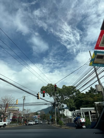 The image is taken from inside a car on a street in San Rafael, Costa Rica. The road is empty except for a motorcycle on the right side and a car in the distance. The sky is blue with scattered white clouds and there are many power lines running across the street. There are two traffic lights with red lights on. There are trees and buildings on either side of the road.  A "McDonald's" sign is visible on the left side of the road.  A person on a motorcycle is waiting at a traffic light.