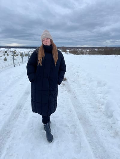 The image depicts a winter landscape, likely in a rural or semi-rural area near Kalinino, Russia. A single young female subject stands on a snow-covered path, centered in the frame and angled slightly left of the camera, exhibiting a subtle smile. She has visible reddish-brown hair beneath a light beige knit beanie. Her attire includes a long, dark (appearing black) quilted puffer coat that reaches mid-calf, dark trousers, and dark ankle boots. Her hands are placed inside her coat pockets.
The path beneath her is compacted snow, showing distinct tire tracks and faint footprints. To the left of the path, a grey metal mesh fence runs parallel, behind which several small, snow-dusted evergreen trees are planted in rows. To the right, an open field is covered in undisturbed snow. The background features a distant treeline consisting of bare deciduous and some evergreen trees, forming the horizon. The sky is uniformly overcast with shades of grey clouds, indicating diffused natural light.