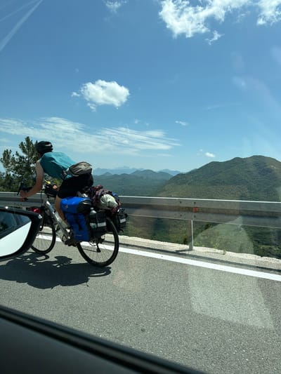 A person on a bicycle with panniers rides along a road on a sunny day in Dobrsko selo, Montenegro. The cyclist, wearing a helmet and a teal shirt, is seen from behind and to the side. The bicycle is equipped with multiple bags, indicating a long journey. In the background, rolling green hills and mountains are visible under a blue sky with scattered clouds. A metal guardrail separates the road from the scenic vista. The image is captured from a moving vehicle, as evidenced by the side-view mirror and the foreground framing.