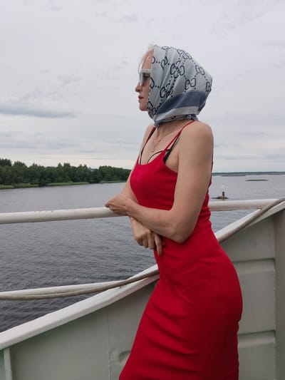 A woman stands on the deck of a ship, looking out at a wide body of water. She is wearing a red, form-fitting dress and a light blue headscarf with a repeating gray pattern. She has on white sunglasses and a delicate silver necklace. Her arms are crossed in front of her as she leans against the ship's railing. The water is choppy and gray, suggesting a cloudy day. In the distance, a line of trees forms the shoreline, and a small lighthouse or buoy stands in the water. The sky is overcast with thick, gray clouds. The overall mood is contemplative, with the woman seemingly lost in thought as she gazes at the horizon. The setting appears to be a river or a large lake in Bereg, Russia. The time of day is difficult to determine due to the cloudy conditions, but it appears to be daytime.