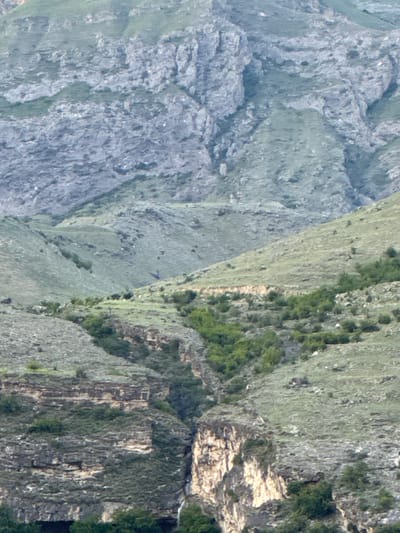 The image depicts a rugged, mountainous landscape with steep, rocky slopes covered in sparse vegetation. In the foreground, a deep ravine cuts through the terrain, revealing layered rock formations. A small waterfall cascades down the side of the ravine. The overall scene suggests a remote, natural environment. The location is identified as Khadzhalmakhi, Russia. There are no people or specific man-made structures visible in the image, only natural geological formations and vegetation.