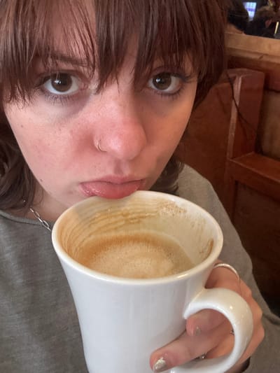 A woman with brown hair and bangs tilts her head, holding a large white mug of coffee. The mug is nearly empty, with coffee remnants visible on the inside. The woman has a nose ring and several rings on her fingers. She's wearing a gray top. A wooden object is visible in the background. The overall impression is casual and intimate.