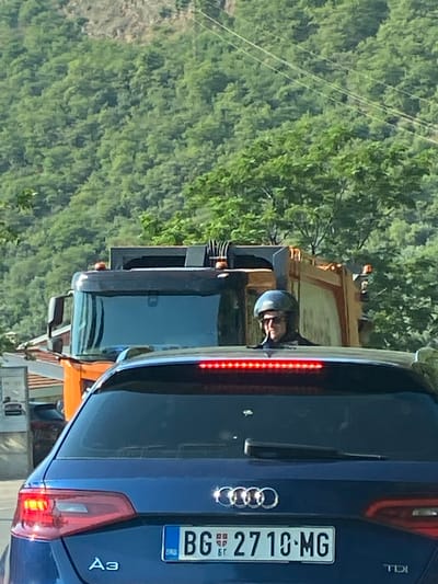 In Donji Katun, Montenegro, on a sunny day, a blue Audi is stopped behind an orange garbage truck. The Audi has a license plate that reads "BG 2710-MG". The driver of the garbage truck is wearing a helmet and sunglasses and appears to be looking forward. The truck has flashing orange lights on its roof. The background is a lush, green hillside covered in trees. The road appears to be a two-lane road.