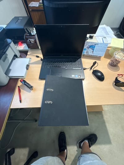 The image displays a top-down view of a cluttered office desk. A black laptop is open, positioned centrally on the desk, with its keyboard visible. To the right of the laptop, a black computer mouse is plugged in. In front of the laptop lies a large, black file folder. To the left of the laptop, a red pen and a black stapler are placed on the desk. A printer is partially visible on the far left, with a white document on its tray. Further to the right, a calendar with the month of June is open, and a clear plastic container filled with nuts sits next to it. A pair of sunglasses rests on a small wooden organizer. The desk is made of light-colored wood, and the background shows office furniture, including a filing cabinet and a computer monitor. The floor is a muted gray. The overall impression is one of a working office environment. The location is stated as the city of Shumentsi, Bulgaria, though there is no visual information in the image to support or refute this.