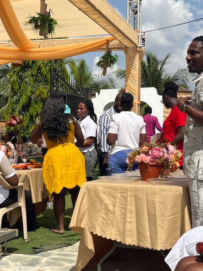 An outdoor social gathering is depicted in a courtyard setting in Kumasi, Ghana, under bright daylight with a partially cloudy sky. The scene is dominated by a large, light-colored canopy structure draped with orange fabric, from which several bamboo-encased artificial plant arrangements are suspended.
In the immediate foreground, to the right, a man with short dark hair, wearing a patterned short-sleeved shirt and a gold wrist watch, is partially visible, facing right and smiling. Directly to his left, a table covered with a light beige scalloped-edge tablecloth holds multiple clear glass charger plates with beaded rims and a brown pot containing a pink and peach artificial floral arrangement.
In the mid-ground, a woman with long, dark, curly hair, wearing a bright yellow, short, textured dress with a scalloped hem and a light blue hair clip, stands with her back to the viewer, barefoot on a patch of artificial green grass. To her left, another woman with braided hair, dressed in a white short-sleeved collared shirt and dark pants, looks towards the left. Behind them, several individuals are mingling, including a man in a striped shirt, a woman in a white ruffled-sleeve top and blue shorts, and a woman in a red top wearing glasses. Further back, a person in a magenta top is visible.
To the far left, another table with a beige scalloped-edge tablecloth displays what appears to be a vibrant orange and blue fabric and clear glasses. A beige plastic chair is also visible. The background features lush green foliage, including palm trees, a black ornate metal gate, and part of a white building with decorative architectural elements. A tall metal truss structure is visible against the sky on the right side of the image. The ground is a mix of artificial green turf and light-colored paving stones, with a rolled-up dark mat or hose on the turf near the barefoot woman.