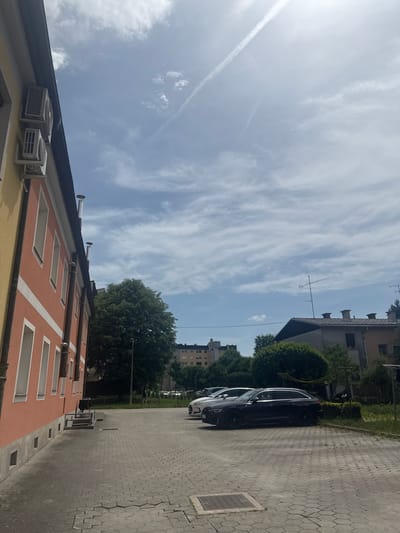 The image shows a street scene in Kranj, Slovenia. The camera is pointed upwards, capturing a view of a building, parked cars, and the sky.
The building in the foreground is light yellow and salmon-colored with a dark roof and white trim around the windows. Air conditioning units are attached to the building. A row of white vent pipes are visible on the roof.
A black car is parked parallel to the building on a paved street. A white car is parked next to it. The street is paved with interlocking gray pavers and a tree is located behind the vehicle.
In the background, a multi-story building is visible behind the tree and beyond the parked cars.
The sky is blue with wispy clouds.