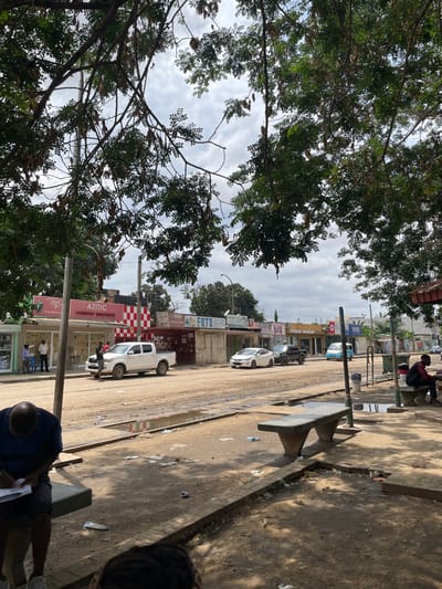This media file captures an outdoor street scene in Luanda, Angola, presented in a portrait orientation but originally captured sideways, depicting a busy street and sidewalk. The sky, partially obscured by lush green trees with dense foliage, suggests an overcast day.
The scene features a street that appears to be unpaved or heavily worn, possibly with a mix of dirt and gravel. Numerous cars, including a prominent white pickup truck and a white sedan, are visible parked or moving along the road. On the right side, a paved sidewalk runs parallel to the street, lined with two concrete benches. One person sits on a bench in the top-right, while another person is seen from an overhead perspective in the bottom-right, seated on a bench and seemingly writing or examining papers.
Buildings line both sides of the street, appearing to be commercial establishments or shops, some with signs. Several other people are scattered along the street and near the buildings, engaged in various daily activities like standing or walking.
Notable details include utility poles running along the street and visible litter, such as plastic bottles, strewn across the sidewalk and road. The overall ambiance suggests a lively, somewhat rugged urban environment.
Visible text on building signs includes "FOTO," "TIGRA," "AZITIC," and "itel," likely indicating businesses or brands present in the area.