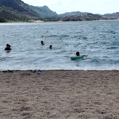 The image depicts a body of water with several people swimming and a sandy beach in the foreground. In the background, a mountainous landscape with sparse vegetation is visible under a partly cloudy sky. On the beach, there are a few scattered rocks and some clothing items. In the water, there are at least five individuals. Two people are in the foreground on the left, with one appearing to be partially submerged and wearing a dark top. Further out, another person's head is visible above the water. To the right of this person, a child is swimming. In the middle ground, a person is swimming with a green inflatable ring. The water appears choppy with waves. The location is identified as being in the city of Торайғыров, Kazakhstan.