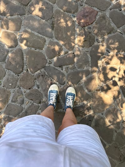 This media file captures an overhead view of a person's legs and feet standing on a cobblestone path. The person is wearing white shorts and blue canvas sneakers with white laces. The cobblestones are varied in size and shape, with visible mortar filling the gaps between them. Sunlight casts dappled shadows from foliage onto the path, suggesting an outdoor setting. The time of day appears to be daytime, likely morning or afternoon, given the quality of the light and shadows. The setting could be a park, garden, or a street in a town. There are no visible emotions conveyed, as the focus is on the lower body. No text is present in the image. The location context of Goryachy Klyuch, Russia, implies this scene might be from a tourist area or a residential street within that region, characterized by similar natural and urban elements.