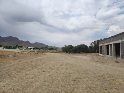 The image depicts an exterior view of an unfinished building under construction in a rural setting, possibly on the outskirts of Egizbulak, Uzbekistan. The foreground is dominated by a dirt road or construction site, with loose gravel and sand. To the right, the unfinished building features cinder block walls, exposed concrete columns, and an incomplete roof structure, suggesting it is in an early stage of construction. In the background, a sparse residential area with low-rise buildings and scattered trees is visible, with a range of mountains forming the distant horizon under a cloudy sky. A yellow pipe runs horizontally across the middle ground, and several concrete barriers are present. The overall impression is one of ongoing development in a somewhat arid environment. There are no people visible in the scene.