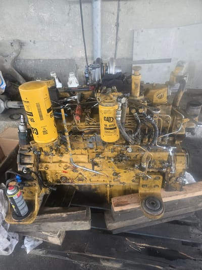 A yellow Caterpillar engine is displayed outdoors, resting on wooden planks. The engine is complex, featuring numerous pipes, filters, and mechanical components. Two prominent yellow cylindrical filters are attached to the engine, one labeled "CAT 1R-0751 Fuel Filter." A can of spray paint is also visible, positioned on a yellow mounting bracket on the left side of the engine. The background shows a concrete wall and a general sense of an industrial or workshop environment. The lighting suggests it is daytime. The location is the city of Galabovo, Bulgaria.