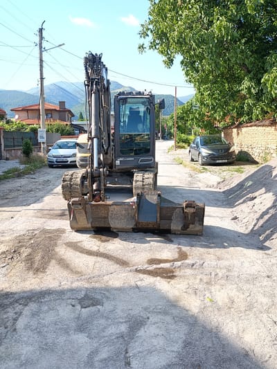 The image depicts an excavator parked on a street in Zlatitsa, Bulgaria. The excavator, a dark grey tracked vehicle with a large bucket, is positioned centrally in the frame, facing forward. Behind the excavator, a paved street extends into the distance, bordered by houses with red-tiled roofs and lush green trees. Two cars are visible parked on the right side of the street. One is a silver hatchback with a license plate reading "CO 0385 CP". The other is a dark grey Peugeot with a license plate reading "CO 7496 BX". A pile of gravel is situated on the right side of the street, adjacent to a brick wall. The sky is clear and blue with a few wispy clouds. The overall scene suggests a construction or road maintenance project is underway.