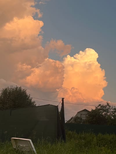 This image depicts a sunset scene in the city of Bolschoi Istok, Russia. The sky is filled with dramatic, orange-tinted clouds, illuminated by the setting sun. In the foreground, tall, green grass grows in front of a dark green fence. A white, plastic chair is overturned in the grass. Behind the fence, a rustic-looking house with a gabled roof is visible, partially obscured by trees. The overall atmosphere is serene and natural, with the vibrant sky dominating the composition.