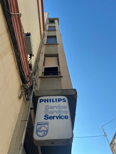 This is a low-angle, eye-level shot of a building's exterior in Castelló De La Plana, Spain. The sky is a clear, bright blue, suggesting a sunny day. The building is made of light-colored stucco and has several windows, some with dark shutters and one with wooden slatted blinds. Electrical conduits and numerous black cables snake across the building's facade, indicating active telecommunications or power infrastructure.
A prominent white sign with blue text and the Philips logo is attached to the side of the building, oriented sideways. The text clearly reads "PHILIPS SERVICE" repeated multiple times. The Philips logo, featuring waves and stars within a circle, is also visible.
On a lower section of the building, a grey electrical box with visible wiring is mounted. The overall scene depicts an urban environment with functional, perhaps slightly aged, infrastructure. The upward perspective and clear sky create a sense of openness, but the abundance of cables suggests a busy, connected location. No people are visible in the frame.