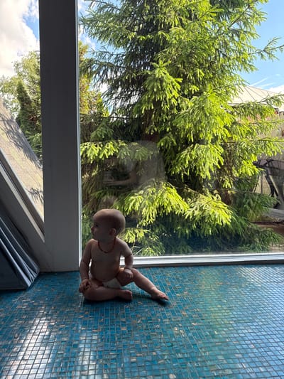 A partially clothed baby sits on a blue-tiled surface near a window. The window shows a dense green tree and sky. The location is likely indoors. The city of Чёрное, Russia, cannot be verified from this image.