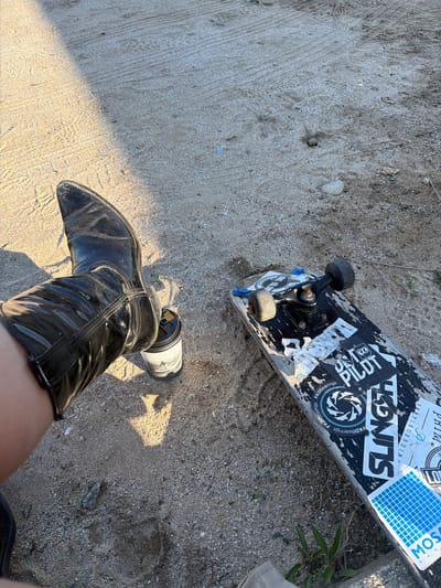 The image depicts a skateboard and a person wearing a black, patent leather cowboy boot on a surface that appears to be a mix of sand and concrete. The skateboard is positioned at an angle to the camera, revealing stickers and graphics on its deck. The boot is pointing towards the lower left corner of the image, and a portion of the wearer's leg is visible. The location is Toseong-myeon, South Korea.