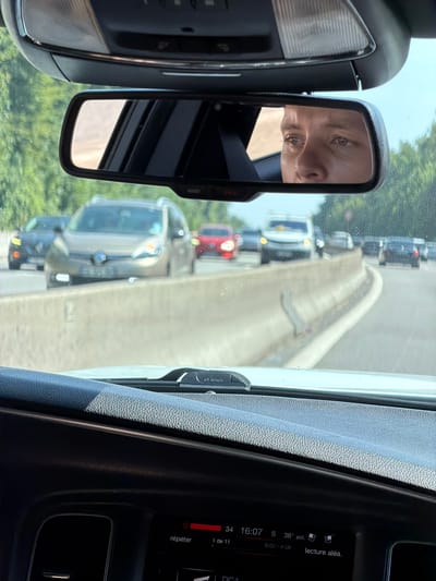 The image is a first-person perspective from inside a vehicle, looking forward through the windshield and upward at the rearview mirror. The rearview mirror reflects the face of a young man, likely the driver, looking forward. The scene outside the car appears to be a traffic jam on a highway, with several cars visible in front, including a light-colored minivan or SUV in the lane to the left. The highway has a concrete barrier. Trees line the side of the road. The dashboard of the car is visible in the foreground, showing a digital display with information such as the date, time (16:07), temperature (38° ext.), and audio playback details. The car appears to be in motion, as indicated by the blurred background and the traffic. The context provided suggests this scene is in Sèvres, France, which would place it within the Île-de-France region.