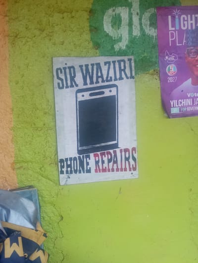 A close-up shot captures a textured, uneven wall painted in vibrant green, with sections of orange and a large green and white "gloo" logo on the upper left. A sign for "SIR WAZIRI PHONE REPAIRS" is centered on the wall, featuring bold blue text and a stylized illustration of a smartphone. To the right, a political poster for "LIGHT PLAY" showcases a man in traditional attire wearing glasses and a cap, with the APC logo and the year 2027 prominently displayed. Below, the text "vote YILCHINI JA FOR GOVERN" is partially visible. On the lower left, a stack of items wrapped in blue and yellow fabric is partially visible, with the letter "M" clearly printed on one of the wrappers.