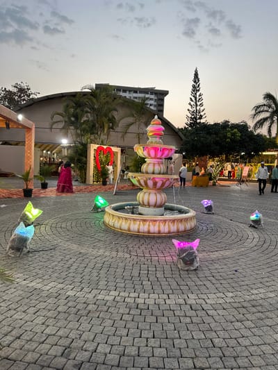 The image shows an outdoor event venue in Nashik, India at twilight. A decorative fountain, illuminated with multicolored lights, is the focal point. People are gathered in the background near decorated structures. The ground is paved with cobblestones. A tall building is visible in the distance.