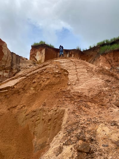 The image depicts a person standing on top of a steep, reddish-brown embankment in Nguzu Edda, Nigeria. The embankment appears to be eroded earth, with visible layers of soil and rock. Sparse green grass grows along the top edge of the embankment. The person is wearing a striped shirt and shorts, and is holding their arms up near their head, possibly taking a picture or posing. The sky above is overcast and cloudy. The overall impression is a rugged, natural landscape with signs of erosion.
