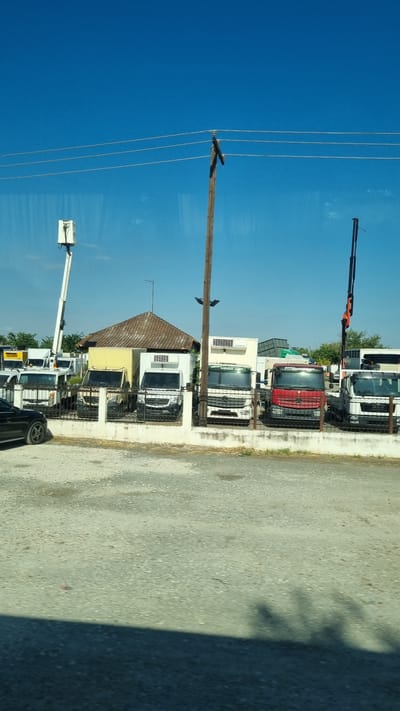 An outdoor scene in the Municipal Unit of Larissa, Greece, captures a storage lot filled with commercial vehicles under a clear blue sky. A wooden utility pole stands prominently in the foreground, with power lines stretching across the frame. Various trucks, including refrigerated vans and a red flatbed truck, are parked behind a chain-link fence. To the left, a white cherry picker extends upwards. A small, dilapidated building with a tiled roof sits behind the parked vehicles. The ground is covered with gravel, and a shadow of foliage falls across the lower portion of the image.