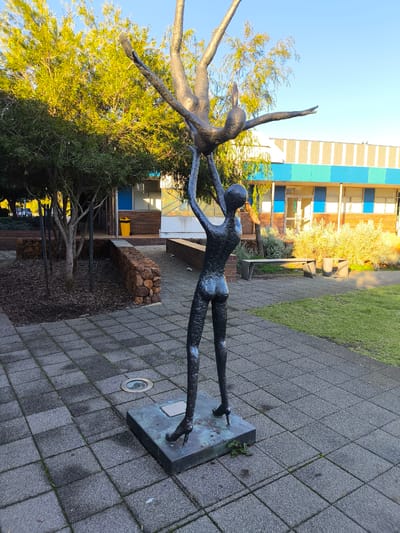 The image depicts a bronze sculpture of two figures in an outdoor setting. The sculpture features one figure being held aloft by another, with both figures in dynamic, stretched poses. The lower figure is standing and appears to be supporting the upper figure, which is positioned upside down. Both figures are rendered in a stylized, elongated form.

The sculpture is situated on a paved area made of square grey tiles. To the left of the sculpture, there is a garden bed with mulch and several small trees with thin trunks and leafy canopies. Behind the sculpture, a low wall constructed of rough stones separates the paved area from the garden bed. Further back, a low-rise building with a brick lower section and a facade featuring horizontal bands of blue and cream colored panels is visible. The building has windows and a glass door with "LOCKIE" signage. In front of the building, there is a seating area with a bench and two cube-shaped seats, surrounded by low-lying green shrubs that are illuminated by sunlight. A yellow trash can is located near the entrance of the building. The sky above is clear and blue. The image was taken in Byford, Australia.