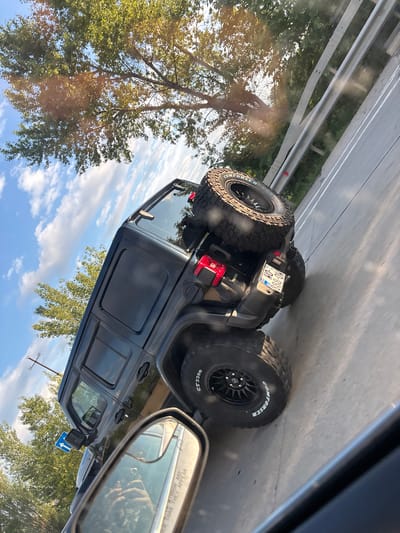 A black Jeep Wrangler is traveling on a highway, likely in Berbuki, Georgia, given the context. The vehicle is viewed from the rear-right side. It has a spare tire mounted on the rear cargo door and custom rims on its wheels. The license plate is visible and appears to be a local Georgia plate with the number "W0-888-GH". The Jeep is on a paved road with a metal guardrail to its right. Trees and a bright blue sky with white clouds are visible in the background, suggesting it is daytime. A power pole is also visible on the left side of the image. The perspective suggests the photo was taken from another vehicle.