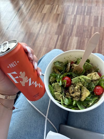 This media file captures a close-up view of a person holding an orange can of "Fever-Tree Ginger Beer" while seated on a surface covered with blue denim, likely jeans. Next to them is a white paper bowl filled with a salad. The salad appears to contain avocado, cherry tomatoes, and leafy greens. A wooden spoon is visible, partially submerged in the salad. The background shows a wooden floor with a parquet pattern.
The image seems to be taken during the daytime, with natural lighting. The setting appears casual and possibly indoors or in a shaded outdoor area, given the lighting and the presence of a laptop on the right side of the frame. The person's hand is adorned with colorful nail art, and they are wearing a watch on their wrist.
Visible text on the can reads "Fever-Tree" in a stylized font, "Ginger Beer," and "PRET*". The overall mood conveyed is one of a relaxed meal or break. While the specific location is given as Orio al Serio, Italy, there are no explicit visual cues within the image to confirm this geographical context.
