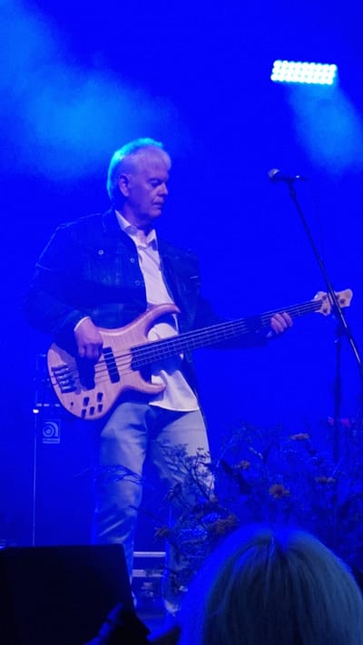 A man with gray hair plays a bass guitar on a dimly lit stage. He is wearing a dark jacket over a white shirt and light-colored jeans. A microphone stands to his right, and a bright light is visible in the background. The overall scene is bathed in a blue light, creating a smoky atmosphere. The image likely depicts a musical performance in Priekule, Latvia.