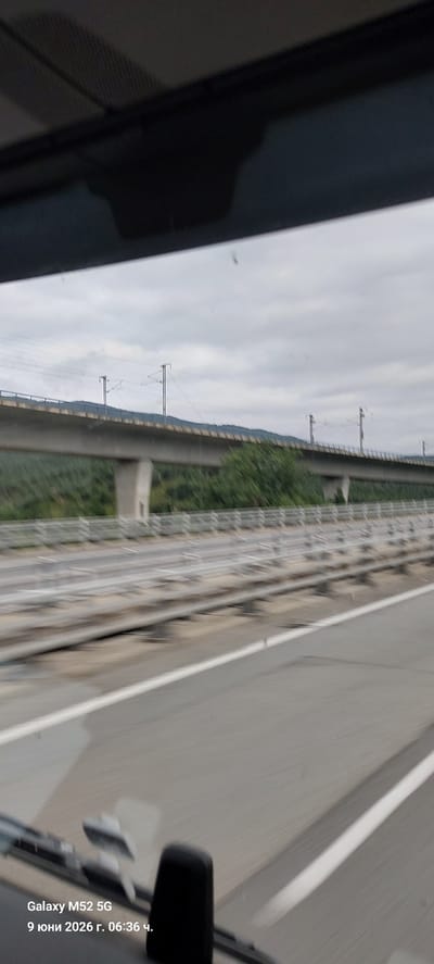 The image shows a view from a vehicle moving along a highway or elevated road. In the background, there is a large concrete overpass, likely carrying a railway line, supported by pillars and surrounded by green trees and hills under a cloudy sky. The foreground shows the road surface with white lane markings, and the interior of the vehicle is partially visible at the bottom, including a dashboard element and a phone displaying "Galaxy M52 5G" and the date "9 юни 2026 г. 06:36 ч." The scene is a perspective of transit through a landscape featuring infrastructure and natural elements. There are no people or specific interactions visible. The location is not explicitly indicated as La Jonquera, Spain within the image itself, but it could be a location near the border given the presence of highway infrastructure.