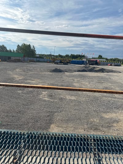 The image depicts an outdoor construction or industrial site. The foreground is dominated by a grated metal barrier, casting shadows on the gravelly ground. Behind this barrier, a large area covered in gravel stretches towards the background. Several piles of gravel are visible, suggesting ongoing construction or material storage.

Further in the background, a blue shipping container and construction equipment, including what appears to be a crane or heavy vehicle, are present. A large truck with a red load bed is also visible, parked near a mound of gravel. Several cars, including a red sedan and a dark-colored SUV, are parked in the middle ground.

A low-rise building with a green roof and a wooden fence is situated on the left side of the image. The sky above is partly cloudy with a mix of blue sky and white clouds. The scene suggests a functional, work-oriented environment. The location is specified as Izyayu, Russia.