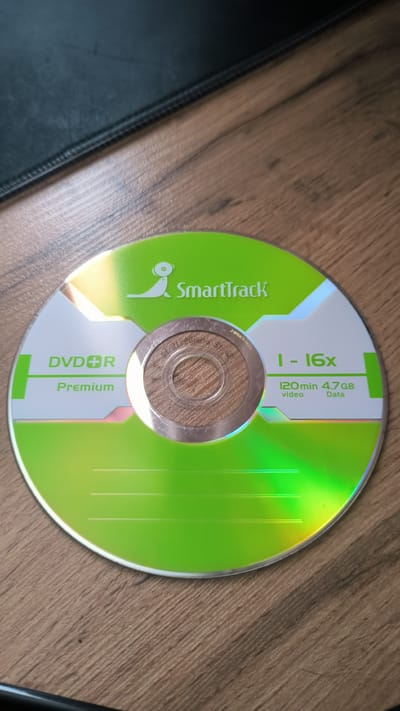 The image shows a DVD-R disc with a bright green and white design. The disc is lying on a wooden surface, likely a table, with a dark, textured object, possibly a mousepad or a book, in the upper left corner. The disc itself has several lines of text and graphics.

In the center of the disc, there's a silver hub with some faint, etched text that is difficult to read. To the left of the hub, the text "DVDR" and below it "Premium" are prominently displayed in white lettering.

To the right of the hub, there are green text elements indicating "1 - 16x". Below this, "120 min" and "4.7GB" are listed, with "video" and "Data" beneath them respectively. A stylized logo featuring what appears to be a leaping figure or animal is positioned above the text "SmartTrack" in green lettering. The overall design is sleek and modern, with the green and white colors dominating the disc's surface. The disc is reflecting light, creating a rainbow effect, suggesting it is under direct lighting. The scene appears to be indoors, given the lighting and the wooden surface. No people or specific events are depicted. The location context of Kovrov, Russia, is not visually evident from the image itself.