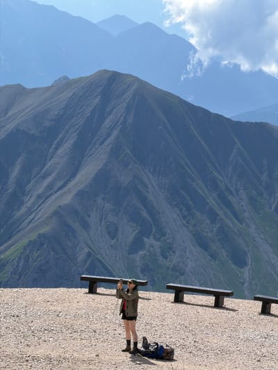A woman stands on a gravelly surface, holding a camera to her face and appearing to take a photograph. She is wearing a green bandana, sunglasses, a green jacket, a red shirt, and black shorts. She has on dark socks and hiking boots. A blue and camouflage backpack lies on the ground near her. In the background, there are several dark wooden benches positioned along the edge of the gravel area. Behind the benches, a vast mountain range dominates the scene, with multiple layers of peaks receding into the distance under a hazy blue sky with scattered clouds. The mountains are rugged, with steep, rocky slopes and patches of green vegetation. The image appears to be taken from a high vantage point, possibly a mountain path or viewpoint. The location is the observation deck of the Mieminger Kette mountain range, specifically on the "Mariahilfberg" overlooking Wildermieming, Austria.