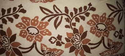 A close-up showcases a floral-patterned fabric. The background is a light beige, adorned with various brown floral and leafy designs. Flowers with round centers and multiple petals are prominent, alternating between lighter and darker shades of brown. Curved stems and detailed leaves weave amongst the blossoms, contributing to a dense and textured overall pattern.