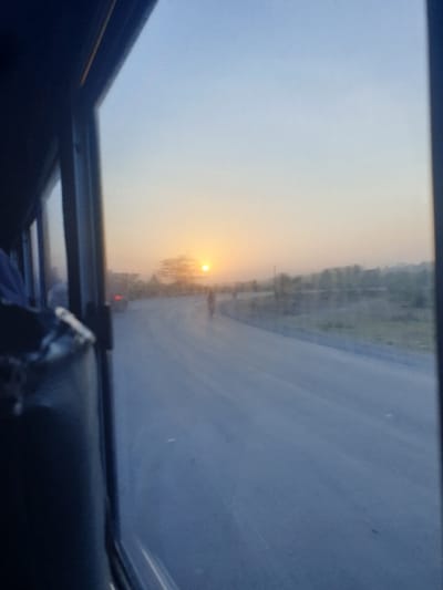 The image captures a hazy sunset viewed through a car's sunroof. The sky is a gradient of blue and orange, with the sun appearing as a bright spot on the horizon. A road stretches towards the sun, flanked by indistinct trees or vegetation. The view is framed by the dark interior of the vehicle, including the sunroof frame and a portion of the car seat.