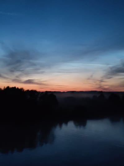 The image depicts a serene, slightly hazy twilight scene over a body of water. The sky transitions from a deep blue at the top to lighter shades of blue and a faint orange and pink hue near the horizon, indicating sunset or sunrise. Silhouetted trees and vegetation form a dark line across the middle ground, with a layer of mist or fog appearing to hug the treeline. The foreground is dominated by the dark, reflective surface of the water, mirroring the sky and the shadowy landscape. No people, specific structures, or identifiable landmarks are visible to suggest a location within the city of Pruzhinino, Russia. The overall impression is one of natural tranquility during a transitional time of day.