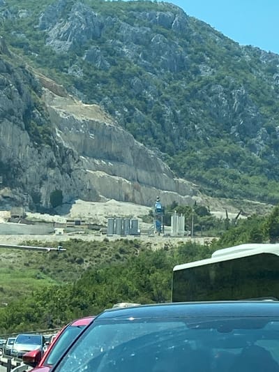 The image captures a mountainside quarry in Mišići, Montenegro, under a clear blue sky. The rocky terrain is extensively terraced, with visible signs of excavation and construction activity. In the mid-ground, several large cylindrical structures, possibly silos, are present. A tall, blue and silver piece of machinery, resembling a construction crane or conveyor system, is positioned near the silos. The lower portion of the image shows a highway with several cars, including a red car in the foreground. Dense green foliage covers the lower slopes of the mountain and the area surrounding the highway. The scene suggests ongoing industrial or infrastructural development in a mountainous coastal region.