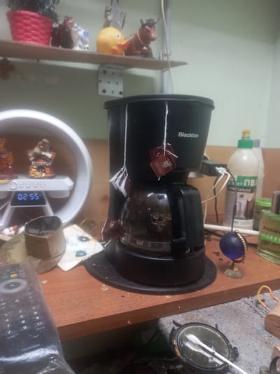 This image depicts a cluttered tabletop in what appears to be a domestic setting. A black coffee maker, identified by the "Blackton" logo, is the central object. Two tea bags are hanging from the coffee maker by strings. To the left of the coffee maker, a white circular device with a digital display showing "02:55" is present, with small figurines on top. To the right of the coffee maker, several bottles are visible, including one with a green cap labeled "ALLIGATOR GREEN." A small blue orb on a stand sits in front of the bottles. In the foreground, a remote control wrapped in plastic is lying on the table. The overall environment suggests a disorganized surface with various items including cleaning supplies and decorative objects. The location in Maloye Lugovoye, Russia is not visually ascertainable from the image.