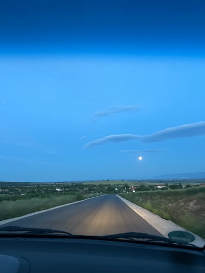 The image shows a road as seen from inside a vehicle, possibly a car, with the windshield visible in the foreground. The road stretches forward and seems to be straight, lit by the vehicle's headlights. Along the road, there is vegetation, including grass and trees, and buildings that suggest it's passing through or near a residential area. The sky above the road is blue with some clouds and a full moon. The angle and view suggest the vehicle is moving forward on the road at night, near the village of Smilets, Bulgaria.