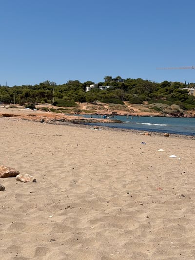 This is a horizontal, slightly low-angle shot of a sandy beach with people in the shallow water. The scene transitions from a clear blue sky on the left to a densely wooded hillside on the right, which slopes down to meet the water. The beach itself is composed of fine, light-brown sand with scattered pebbles and small shells. A narrow strip of rocks and rough terrain separates the sand from the water's edge.
Several people are visible in the water, appearing to be swimming or wading. A few more figures can be seen on the hillside, perhaps walking or relaxing. The vegetation on the hillside is thick and green, with some areas of bare earth and rocks visible. In the distance, atop the hill, there are glimpses of white buildings. A tall construction crane is visible in the upper left portion of the frame, silhouetted against the sky, suggesting ongoing development or construction in the area.
The water is a bright turquoise and appears calm, with small waves lapping at the shore. The lighting suggests it is a sunny day, likely late morning or early afternoon, casting sharp shadows. The overall mood is one of leisure and recreation, with people enjoying a day at the beach. The location appears to be a Mediterranean coastal area, consistent with Vouliagmeni, Greece. There is no visible text in the image.