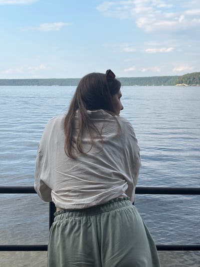 A woman stands on a walkway, her back to the camera, facing a wide body of water. She has long, dark brown hair and is wearing a loose, light-colored shirt and pale green pants. Her head is turned to the right, and she appears to be looking out at the water. Sunglasses are perched on top of her head. The water stretches out to a tree-lined horizon, with a pale blue sky with scattered clouds above. The location is the shore of a large lake or river in Lyady, Russia.