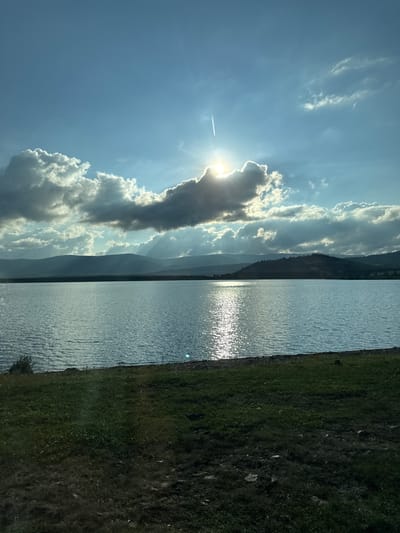 The image depicts a serene natural landscape, likely a reservoir or large lake, under a partially cloudy sky. The sun is shining brightly, partially obscured by a large, dark cloud, creating a dramatic effect with rays of light filtering through. A contrail from an airplane is visible in the sky. The water is calm with a shimmering reflection of the sun on its surface. In the background, rolling hills or mountains form a dark silhouette against the sky. The foreground consists of a grassy embankment sloping down to the water's edge. There are no people or man-made structures visible in the image, suggesting a remote or natural setting. The scene evokes a sense of peace and tranquility. The specific location in the city of Геологоразведка, Russia is not identifiable from the visual information provided in the image itself.