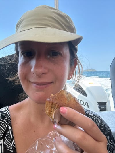 A female individual is depicted in a close-up, chest-up shot aboard a vessel. She wears a beige wide-brimmed sun hat and a black and white patterned top. Her face shows a slight smile, with a small white smudge present below her right eye. She holds a partially consumed, light brown muffin, wrapped at its base in clear plastic, in her left hand, which exhibits pink-painted fingernails. In the midground to her right, a white Honda 250 horsepower outboard motor is visible. The background reveals choppy blue water leading to a distant, hazy landmass under a clear, bright blue sky. The scene is consistent with a coastal or marine environment, specifically in the city of Pak Nam, Thailand.