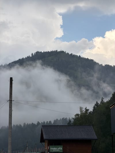 The image depicts a misty mountain landscape in Uznezya, Russia. A thick layer of fog partially obscures a forested mountain, creating a dramatic atmospheric effect. In the foreground, a wooden utility pole with wires stretches across the frame. Below the fog line, a dark-roofed wooden structure with a sign that reads "ТРАВА-МЁД" (Grass-Honey) is visible. The sky above is a mix of clouds and patches of blue. The overall scene conveys a sense of natural beauty and tranquility, with a touch of rustic charm from the foreground structures.